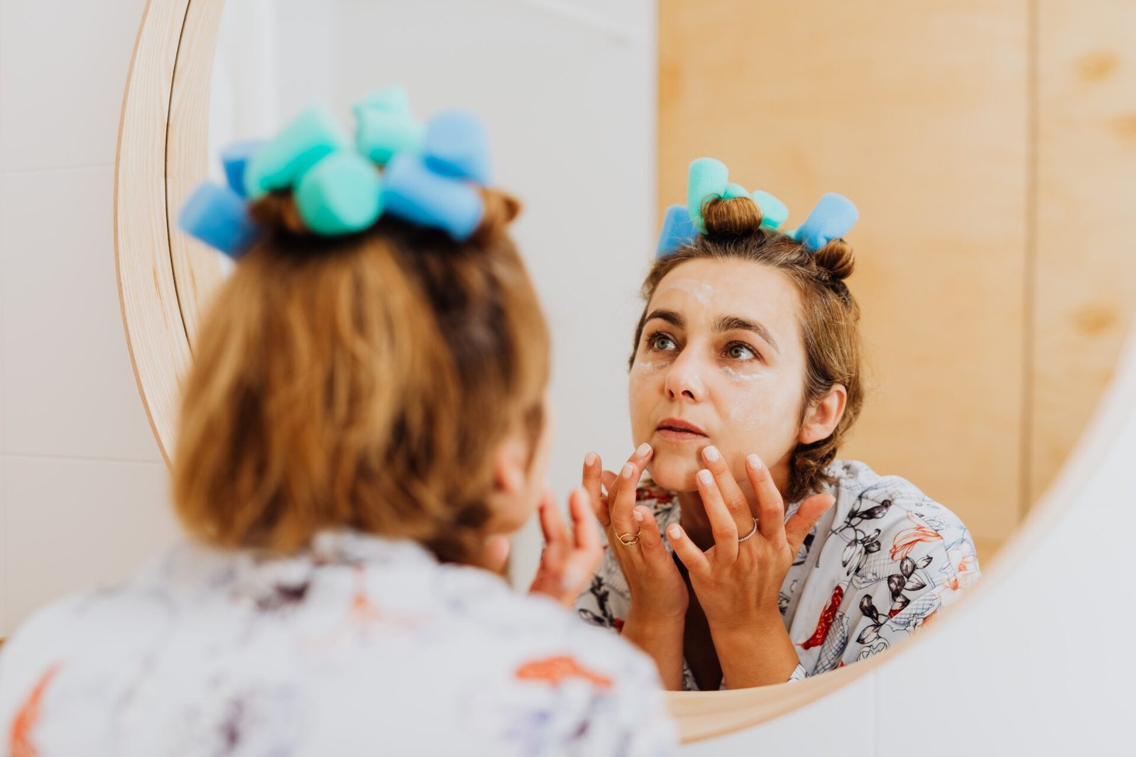 A woman with hair rollers is applying cream to her face while looking at herself in a mirror