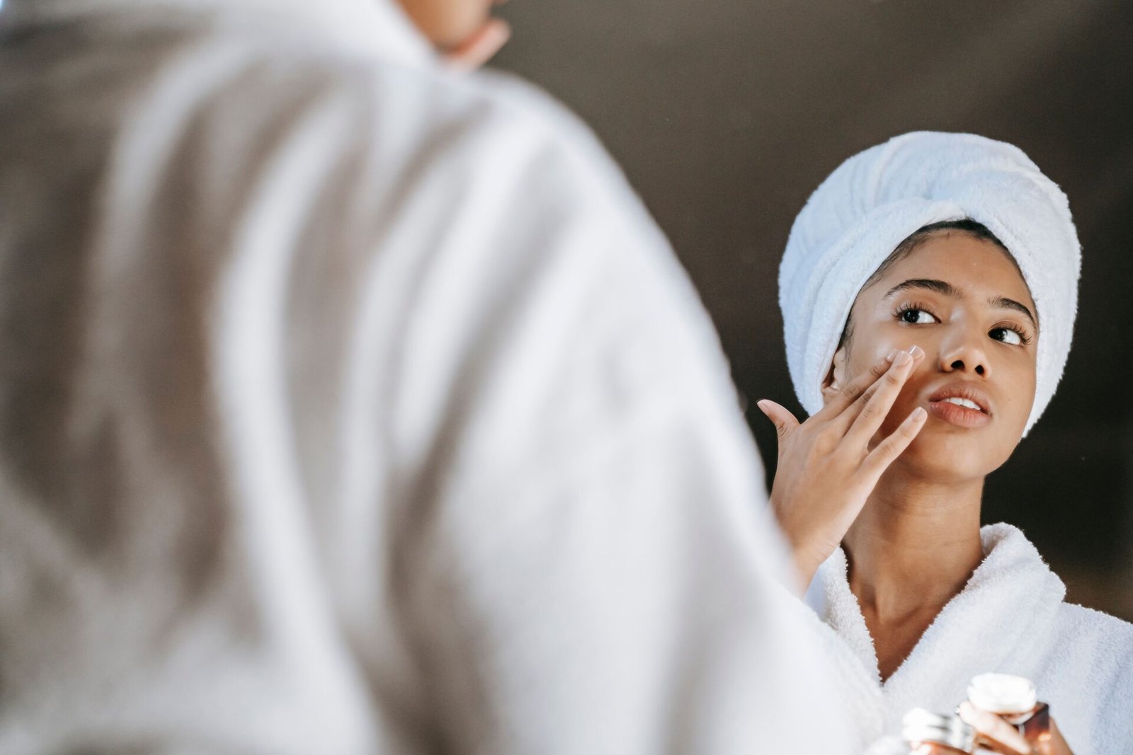 A person in a white robe and towel is applying skincare products while looking at their reflection in a mirror