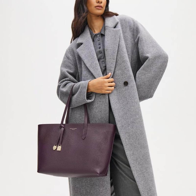 Woman wearing Aspinal of London fig pebble leather East West tote bag with gold shield charm styled with a grey coat.