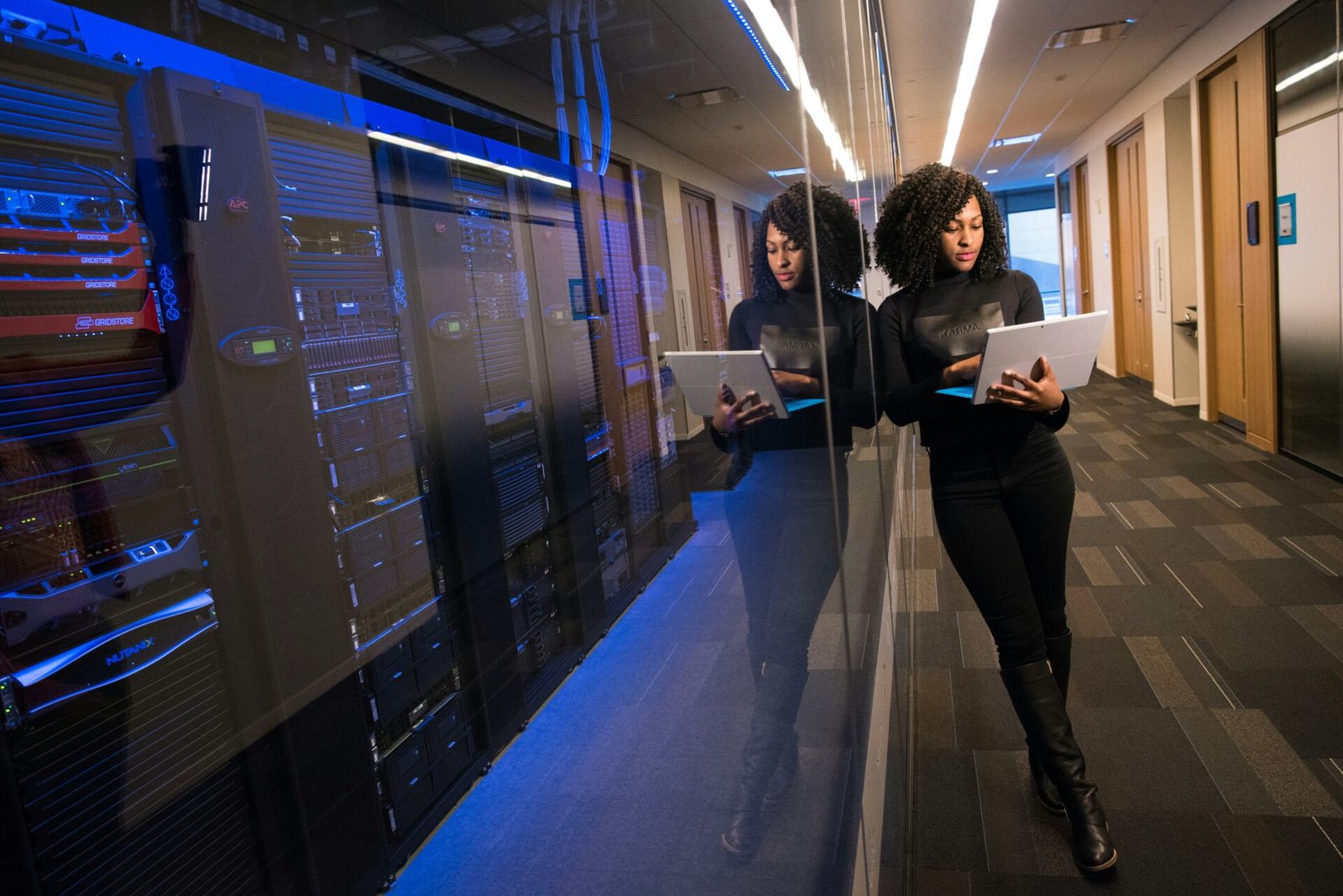 Woman standing in a hallway holding a silver laptop and leaning against a large computer, her reflection is mirrored in the glass