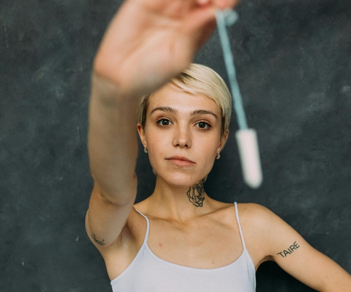 A woman with short blonde hair and taire tattoo on arm holds up a tampon towards the camera against a dark background, highlighting menstrual health and period poverty awareness as Scotland become first country to make free period products for all women.