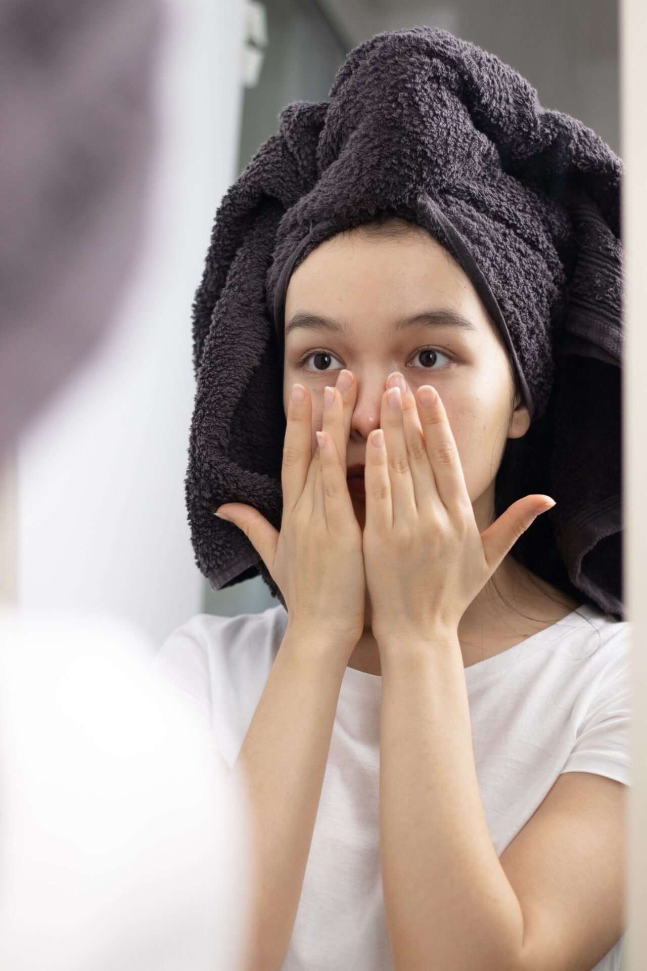 A person with a towel on their head is applying skincare to their face while looking in a mirror