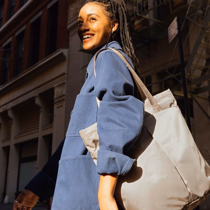 Woman carrying a fawn Bellroy Laneway Totepack styled as a shoulder tote in an urban setting. The versatile design features dual carry options and a spacious interior ideal for commuting or travel.