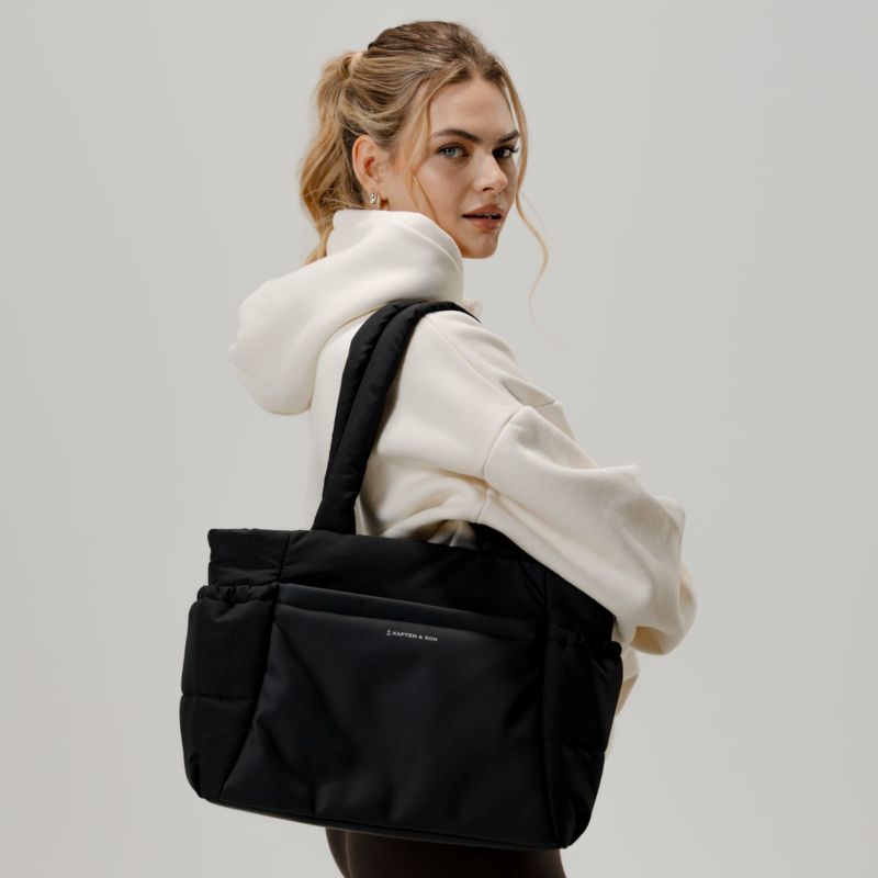 Woman wearing a black Kapten & Son Hellvi Cloud Medium tote bag over a cream hoodie. The padded design and front zip pocket highlight its lightweight, modern everyday style.