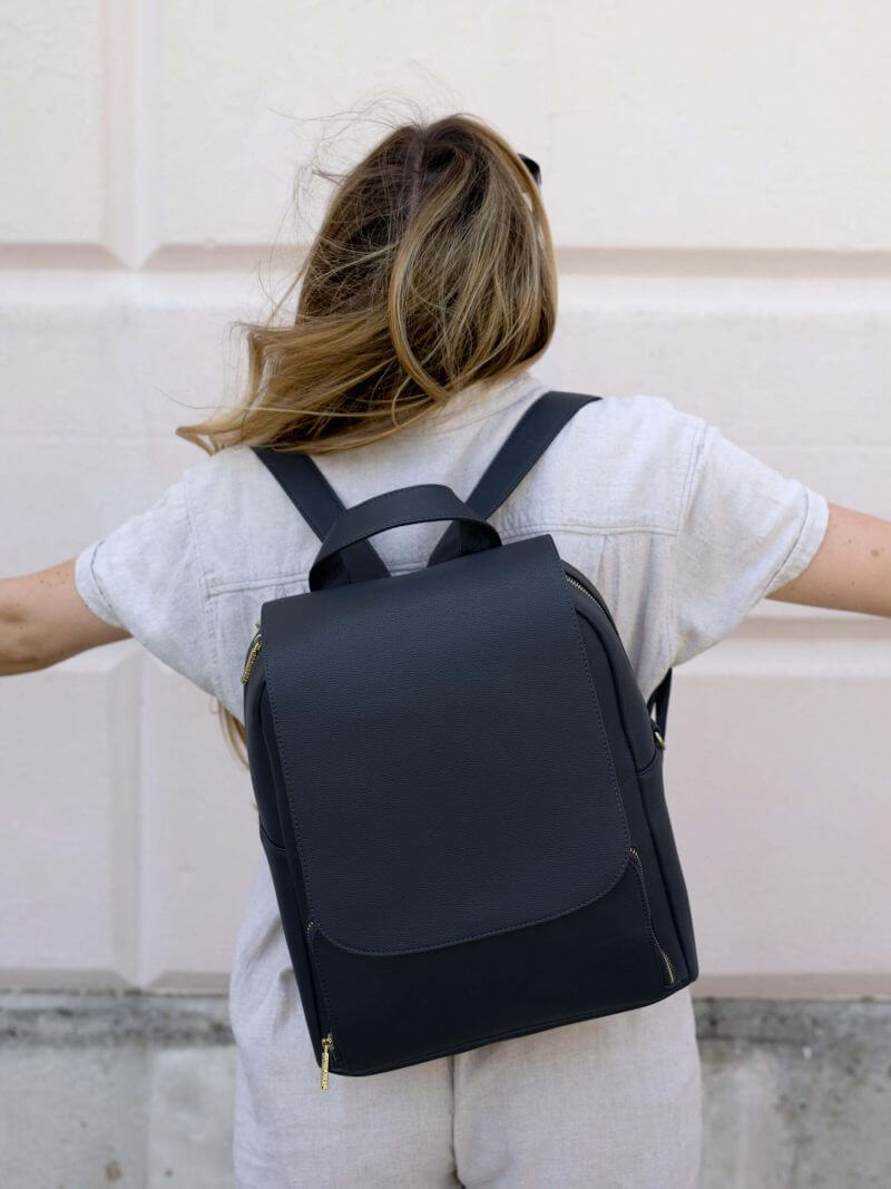 Woman wearing Stackers navy blue backpack with structured flap front, gold zip details and adjustable shoulder straps.