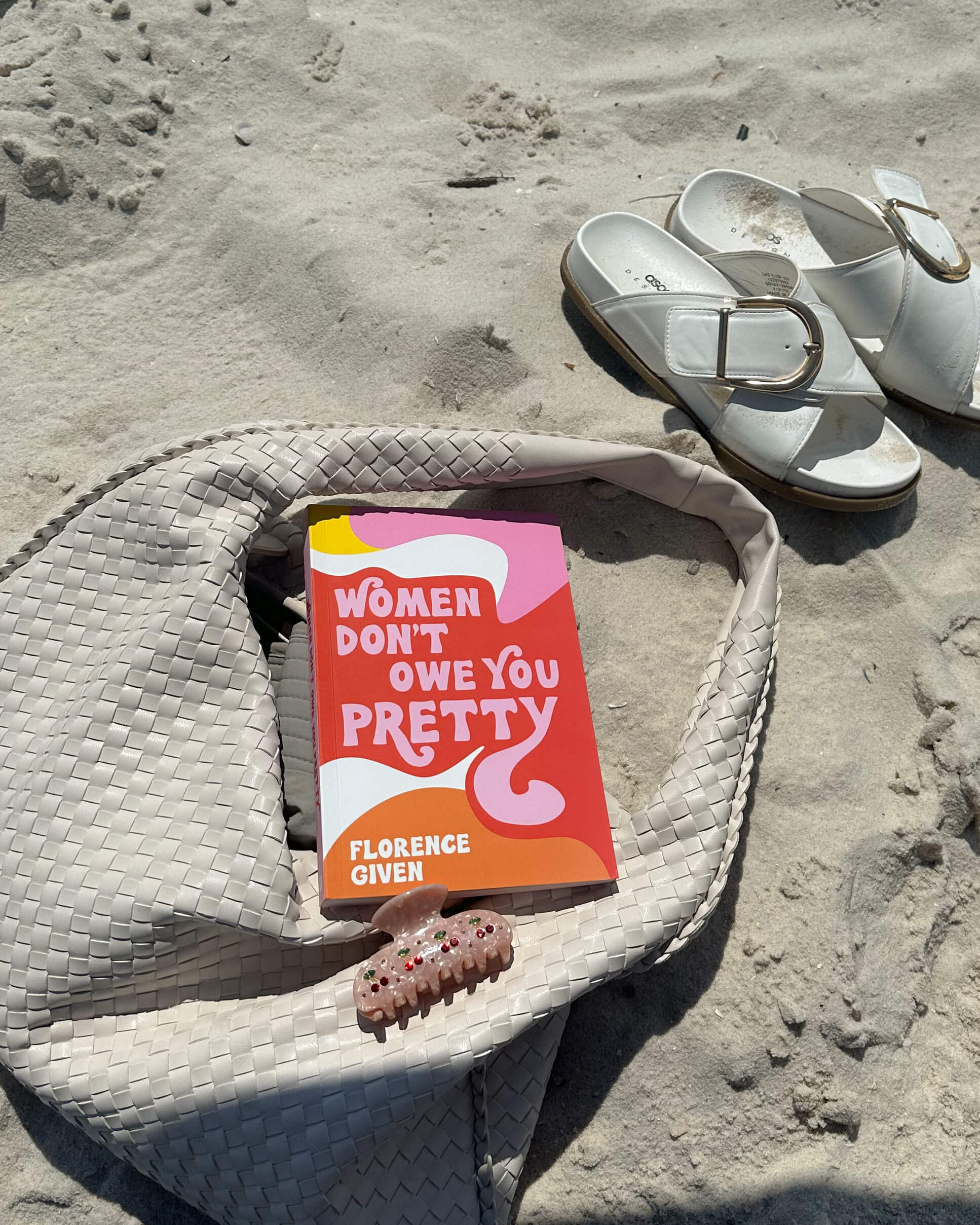 'Women Don't Owe You Pretty' book by Florence Given resting on a woven bag on the beach beside white sandals, styled as a modern feminist summer reading pick - best feminist books