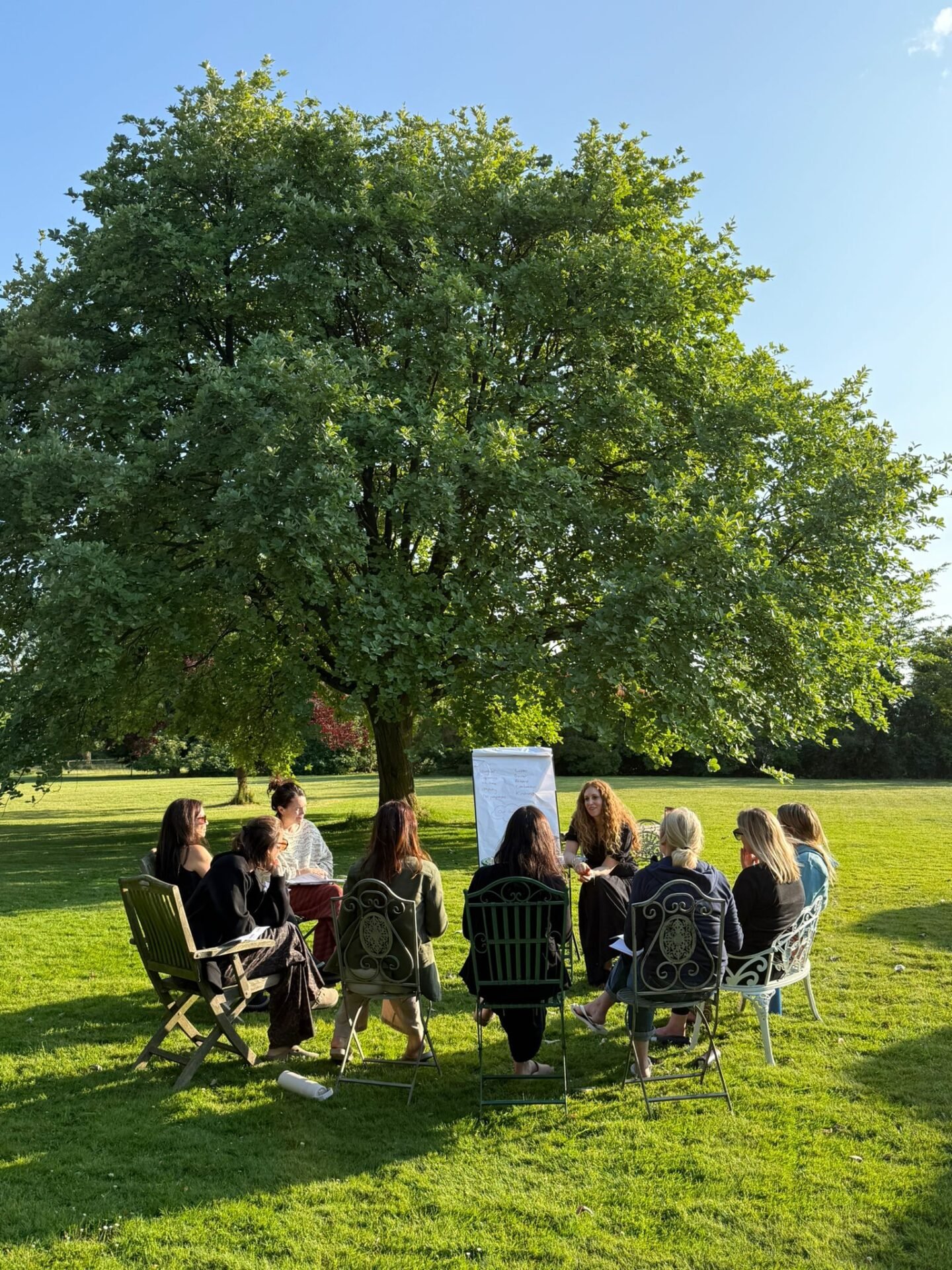 At women-only retreat, Revive & Rise, a group of women sit in a circle on chairs on a grassy lawn under a large, leafy tree, facing a whiteboard during an outdoor workshop session - women-only retreat